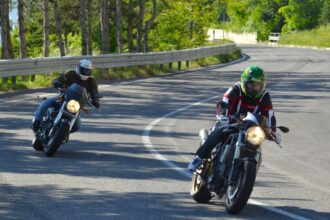 Két motoros kanyarodik a napfényes úton, élvezve a sebességet.