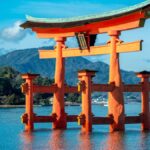 Egy japán torii kapu áll a vízben, háttérben hegyekkel és fákkal.
