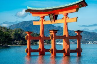 Egy japán torii kapu áll a vízben, háttérben hegyekkel és fákkal.