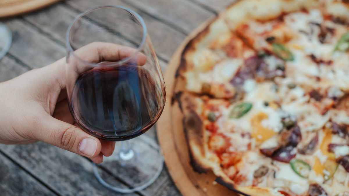 Egy kéz vörösbort tart egy pohárban, mellette pizza látható.